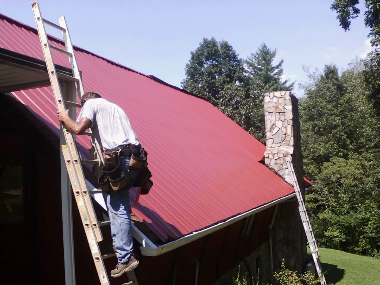 Roofer on a ladder beside a red metal roof and stone chimney — Soffit & Fascia Repair by Roofing Brothers in Westmont